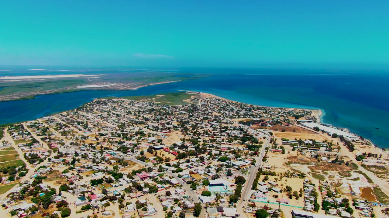 High aerial view over San Carlos, Baja California Sur with residential blocks, sandy beach, turquoise shoreline water, darker ocean beyond, and no visible people.