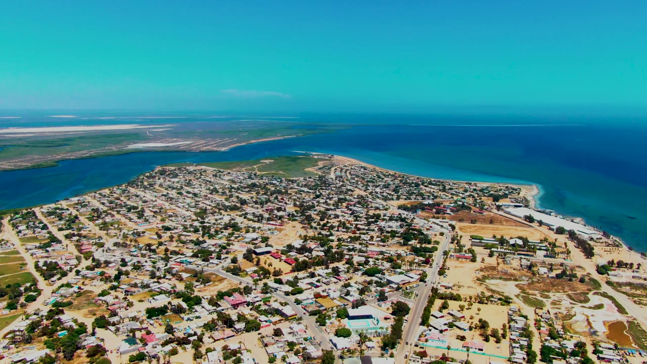 Drone view across rooftops and roads toward a curving beach and ocean.