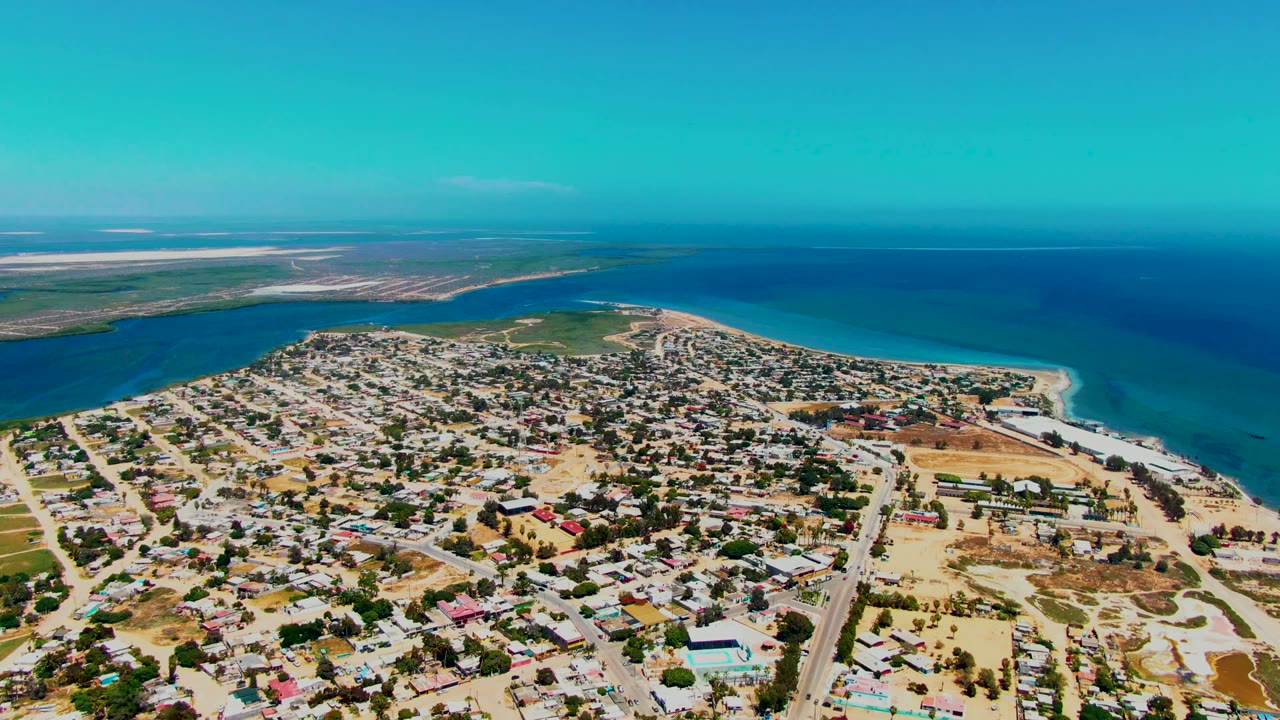 San Carlos appears from above with dense low buildings, straight streets, dry open lots, a bright beach strip, turquoise shallows, and no visible people.