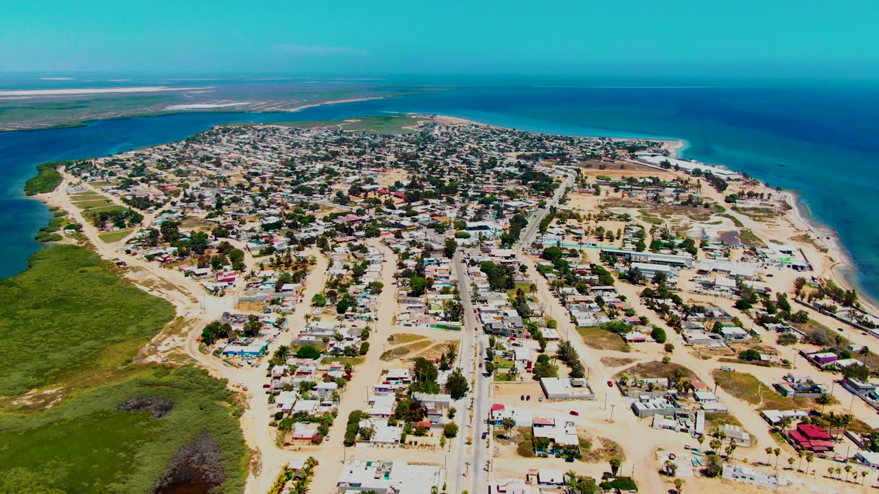 Aerial frame over San Carlos, Baja California Sur, showing sandy streets, low-rise homes, green scrubland, bright turquoise water, and no visible people.