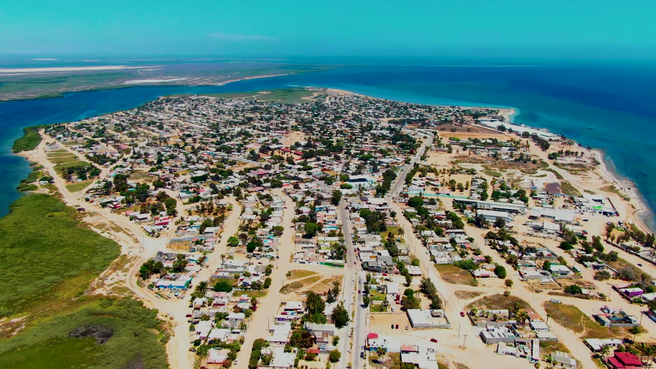High aerial frame of San Carlos showing dense low-rise housing, pale sandy roads, a beach curve on the right, green land on the left, and no visible people.