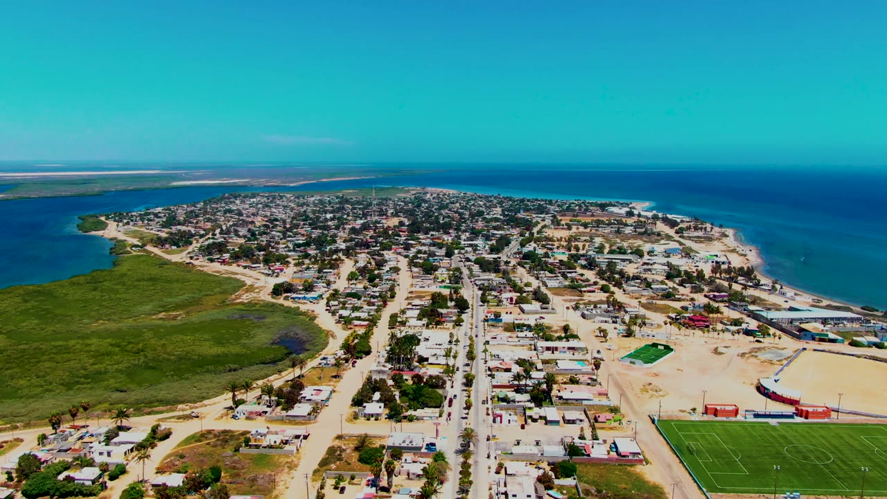 Opening aerial frame over San Carlos, Baja California Sur, with sandy streets, low houses, marshland at left, ocean at right, and no visible people.