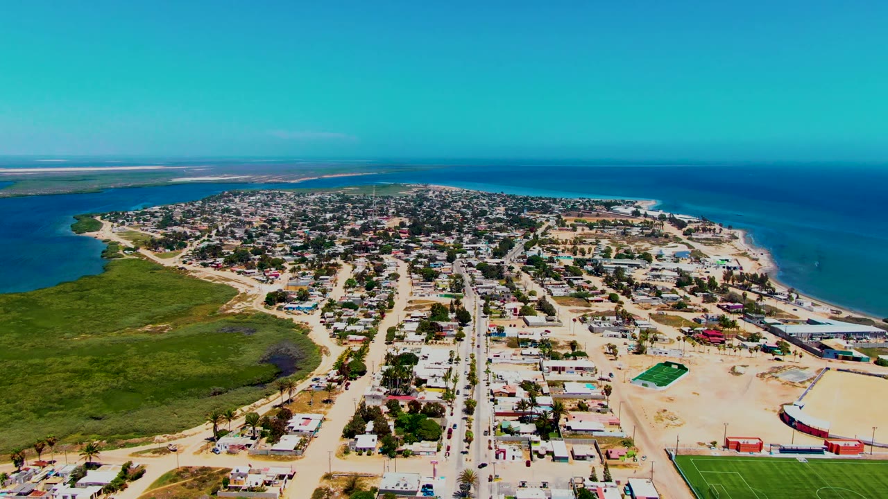 San Carlos appears from above with clustered houses, sandy blocks, a green field near the lower right, marshland to the left, ocean to the right, and no visible people.