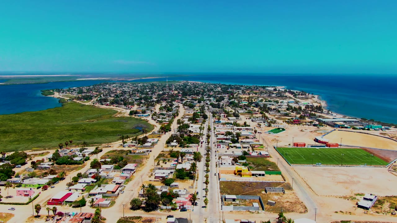 Opening aerial frame over San Carlos, Baja California Sur showing a straight main road through low buildings, a green sports field at right, wetlands at left, blue ocean beyond, and no visible people.