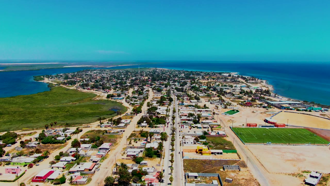 High drone view over a sandy coastal neighborhood, road, green field, and blue water.