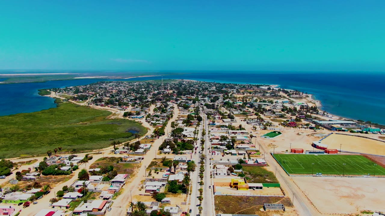Third frame shows San Carlos from above with the green sports field prominent at lower right, a broad sandy area beside it, clustered buildings along the road, blue ocean in back, and no visible people.
