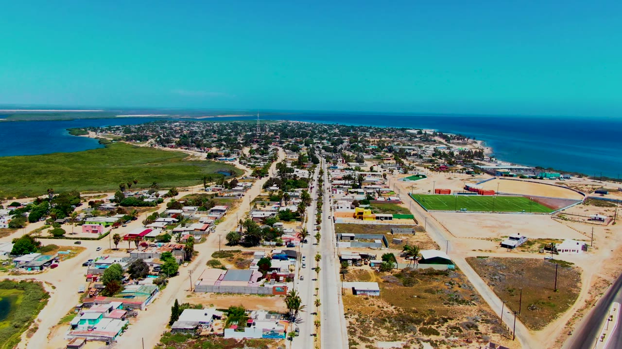 High aerial frame over San Carlos, Baja California Sur, with sparse road traffic, compact homes, a sandy beach edge, green coastal land, open ocean, and no visible people.