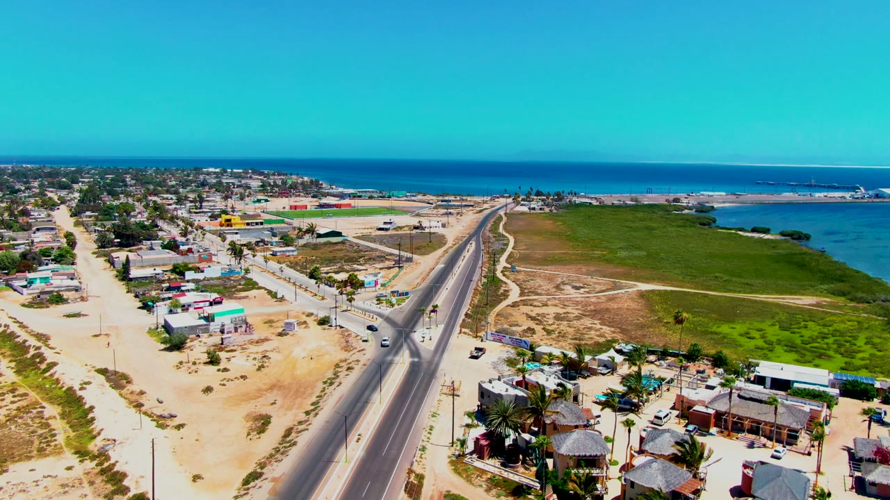 Aerial frame over San Carlos, Baja California Sur shows a straight highway with cars, sandy roadside buildings to the left, green coastal flats to the right, blue ocean beyond, and no visible people.