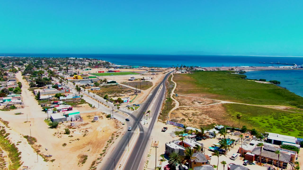 Drone view along a paved road with vehicles, low buildings, open green land, and ocean horizon.