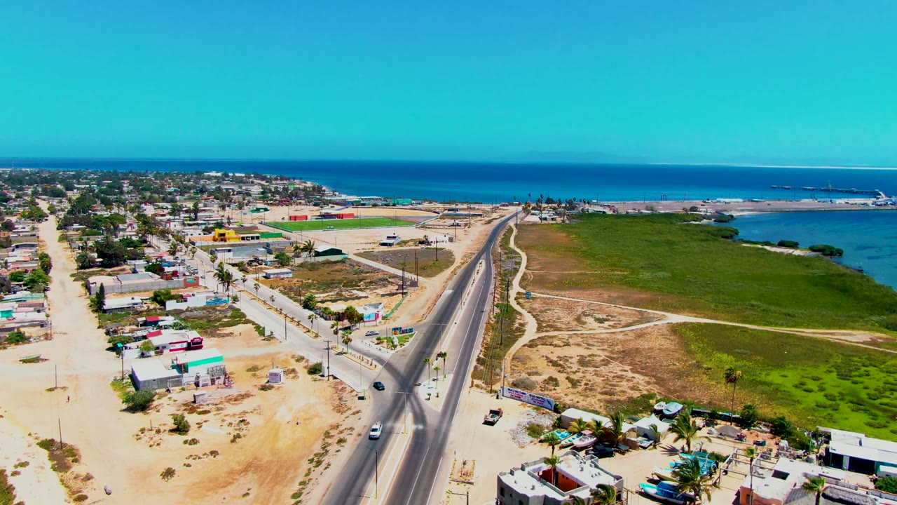 At 0:02, the drone view in San Carlos shows the highway leading toward the coast, with small vehicles, compact buildings, sandy lots, green shoreline land, and no visible people.