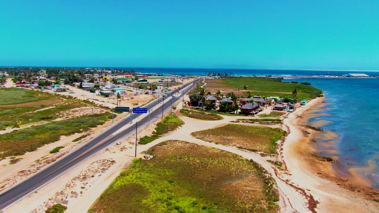 Aerial view in San Carlos, Baja California Sur showing a paved coastal road, sandy beach, small buildings, green fields, blue water, and no visible people.