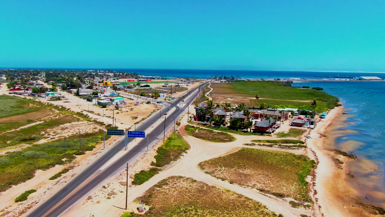 Aerial frame in San Carlos, Baja California Sur showing road signs over the coastal road, clustered small houses, sandy shoreline, shallow blue water, and no visible people.