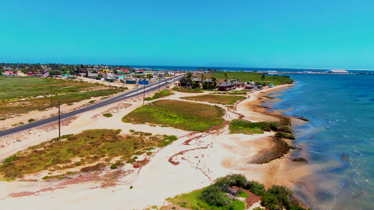 Aerial view in San Carlos, Baja California Sur, showing a coastal road, pale sand, scrub vegetation, blue ocean water, distant buildings, road signs, and no visible people.