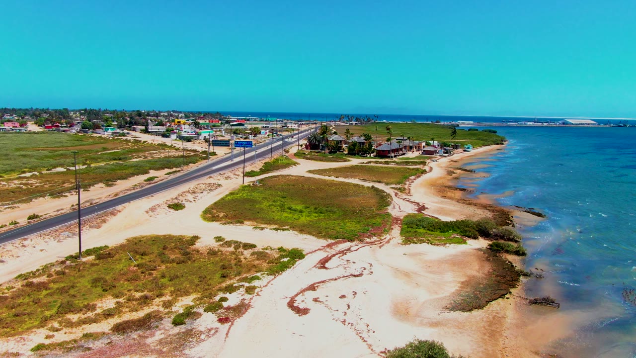 Coastal highway with sandy shoulder, green scrub, ocean, and low buildings.