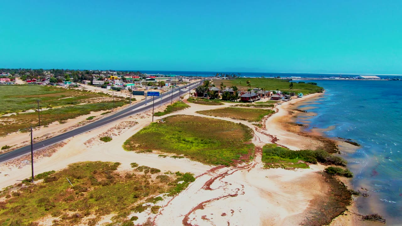 Aerial view in San Carlos, Baja California Sur, showing two road signs near the coastal road, sandy beach curves, green vegetation, blue water, distant buildings, and no visible people.