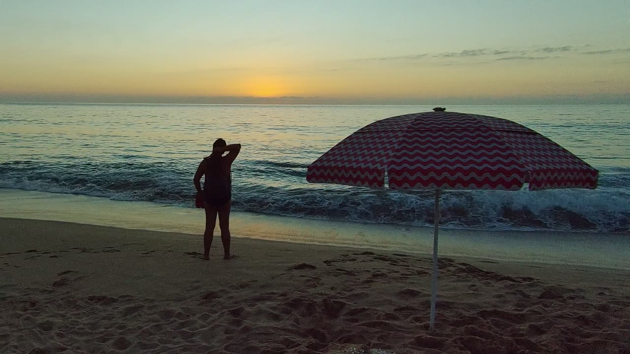 In San Pancho, Nayarit, Mexico, a silhouetted woman stands left of a red and white striped umbrella as small waves reach the sandy shore at sunset.