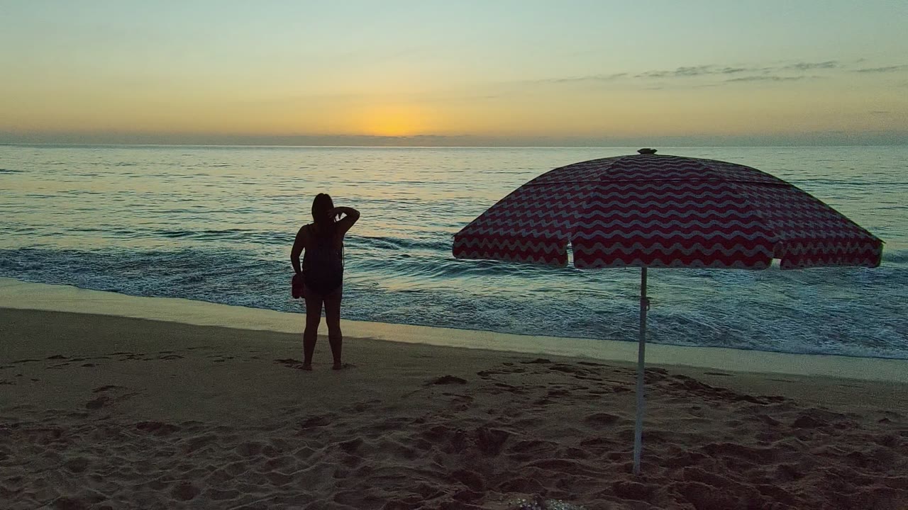 Silhouetted woman adjusts her hair near the shoreline with umbrella at right.
