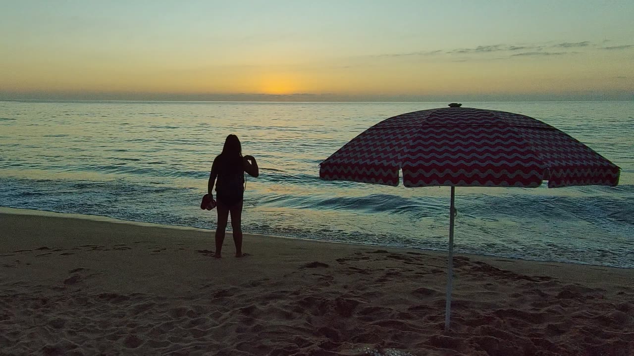 In San Pancho, Nayarit, Mexico, the woman lowers her arm slightly near the waterline while the red and white umbrella covers much of the right foreground under the fading sky.
