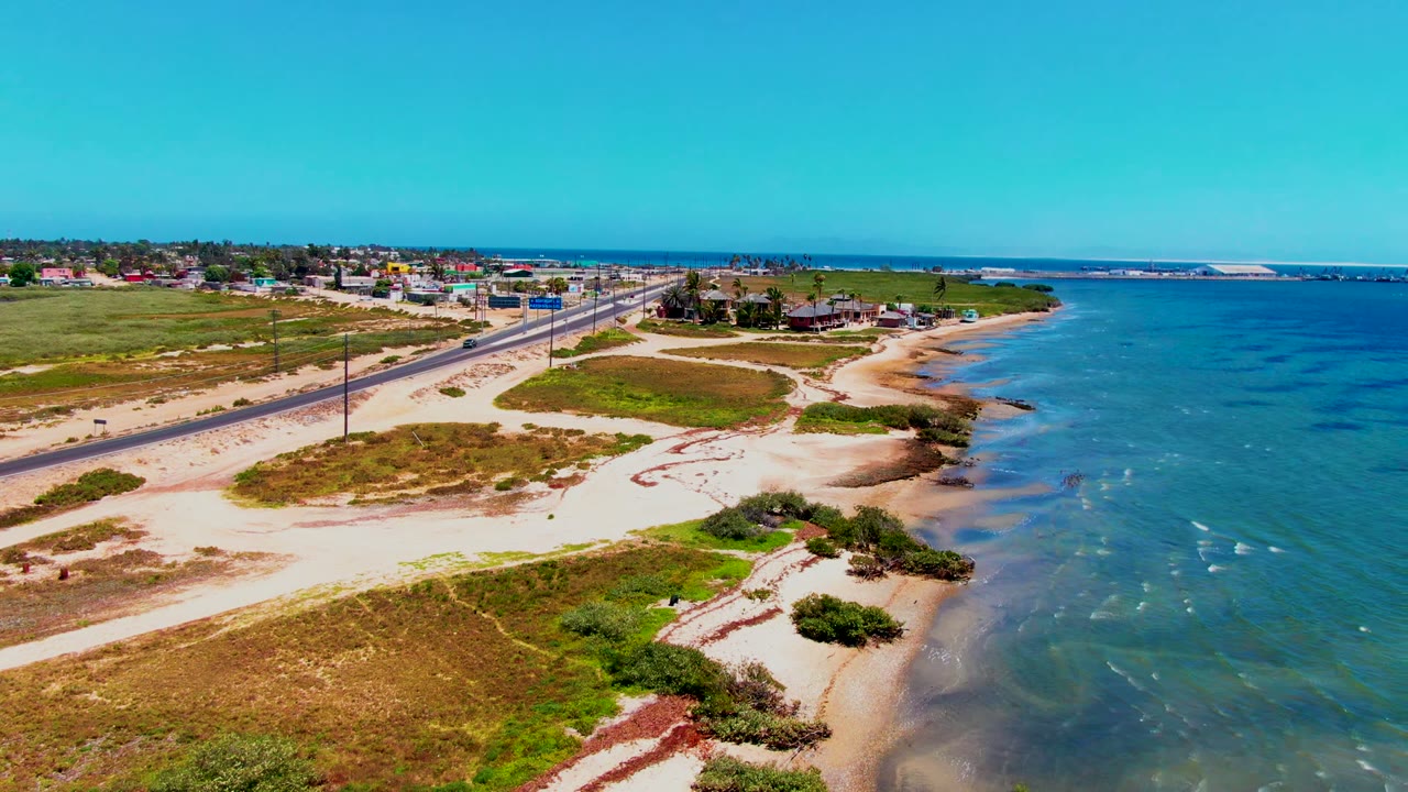 Aerial frame over San Carlos, Baja California Sur showing a paved road beside sandy beach, green dunes, scattered buildings, blue ocean, and no visible people.