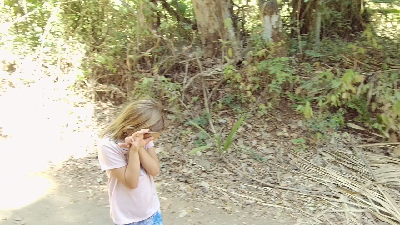 A young girl in a pale pink shirt covers her face near the edge of a dirt forest trail in San Pancho, Nayarit, Mexico, with dry leaves and green brush behind her.