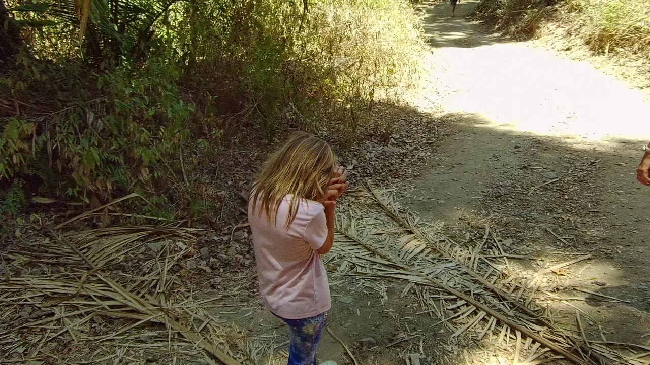 The girl stands on the shaded dirt trail in San Pancho, Nayarit, Mexico, with her hands near her face while part of an adult appears at the right edge of the frame.