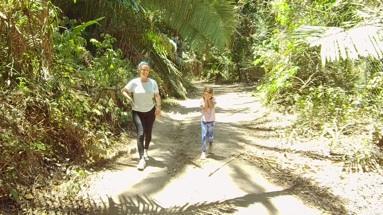 In San Pancho, Mexico, a woman in a light blue shirt and a young girl in a pale shirt walk toward the camera on a sunlit dirt trail bordered by dense foliage.
