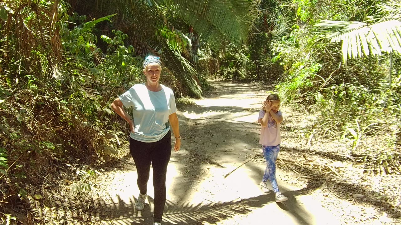 In San Pancho, Mexico, the woman has moved closer on the left side of the frame while the young girl walks to the right, with bright leaf shadows crossing the sandy path.