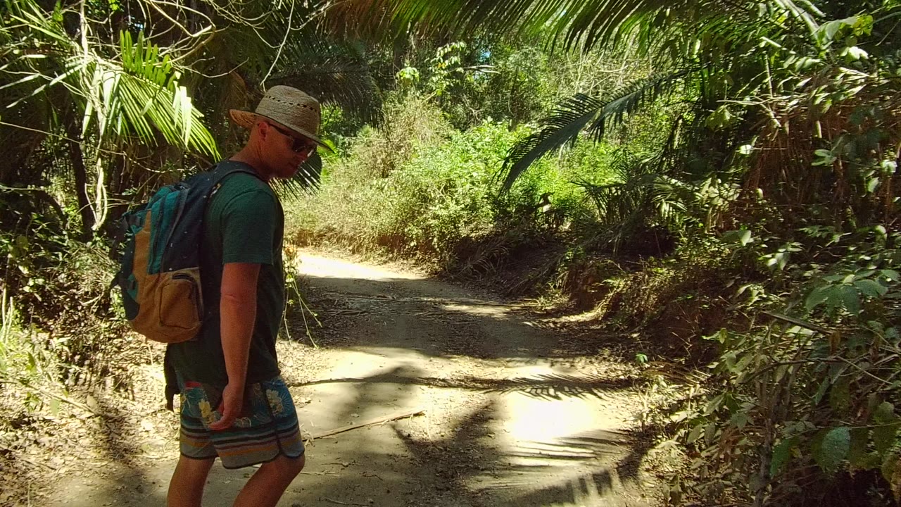 The hiker stands closer to the camera on the right side of the San Pancho dirt path, with bright sun on the trail and dense foliage filling the background.