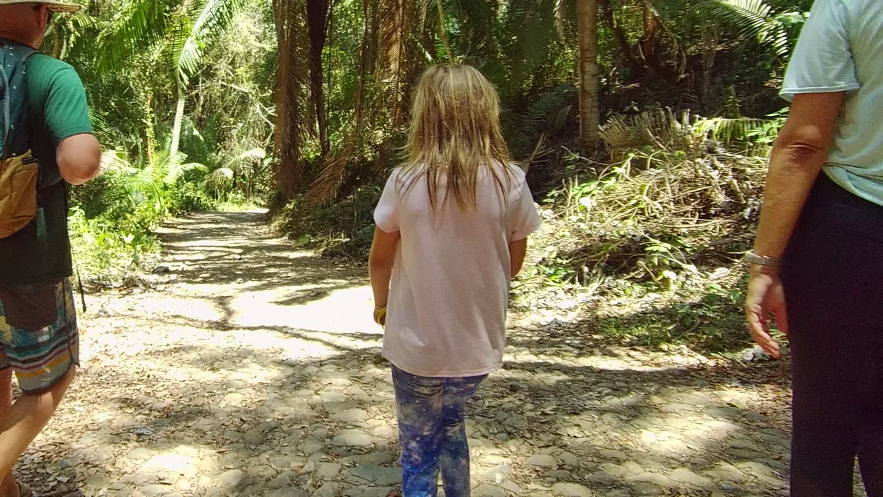A child in a pale shirt walks away on a dirt forest path in San Pancho, Nayarit, Mexico, with adults partly visible at both edges and sunlight on the trail.