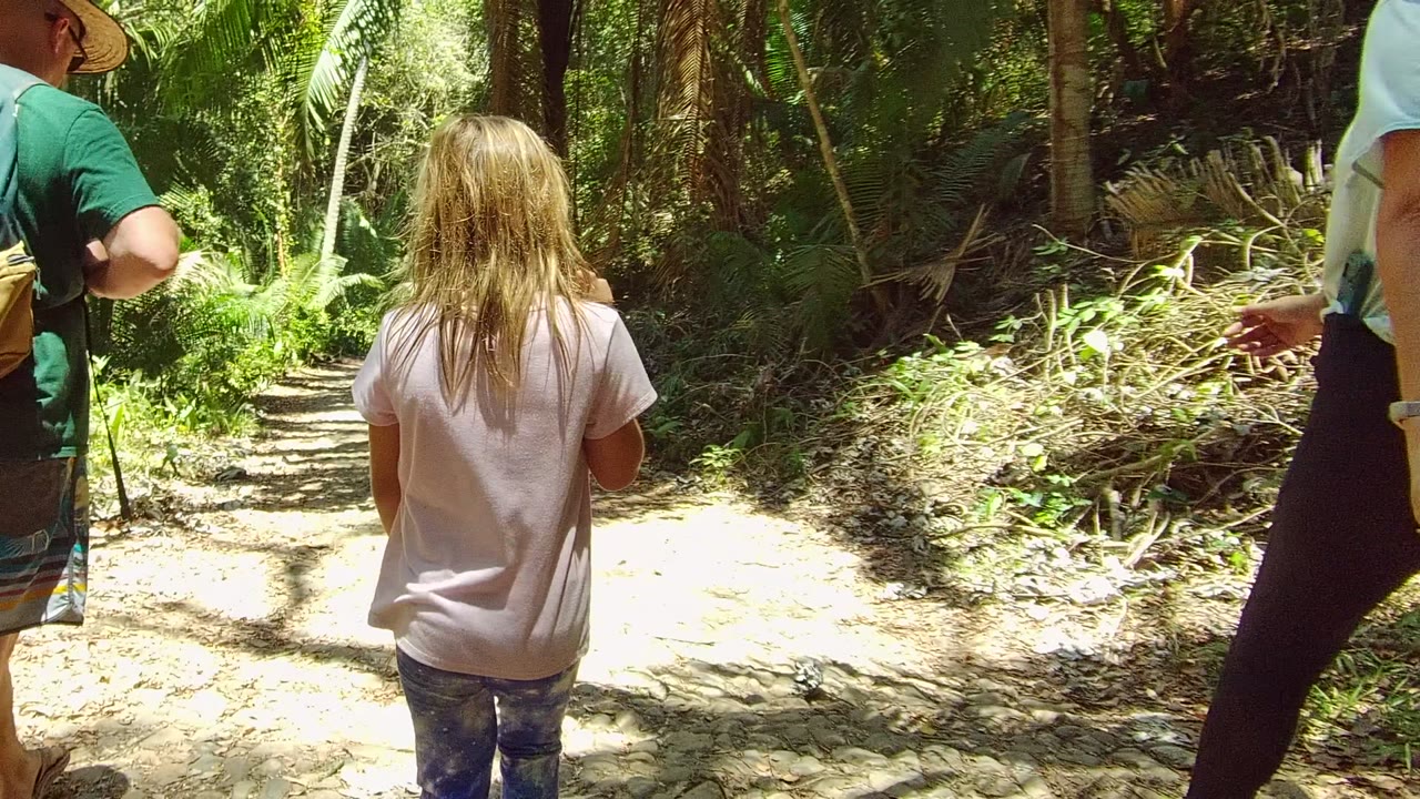 A child walks between two partly cropped adults on a dirt path in San Pancho, Nayarit, Mexico, with thick foliage and bright sunlight breaking through the canopy.