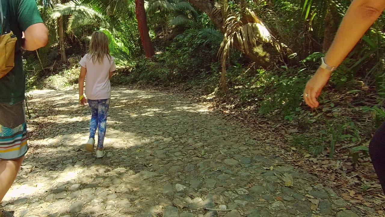 A child walks away along a rocky shaded path in San Pancho, Nayarit, with two adults partly visible at the left and right edges and dense greenery ahead.