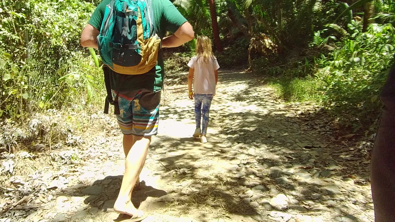 A close foreground arm crosses the right side while the child and backpacked adult remain ahead on the San Pancho dirt trail through thick greenery.