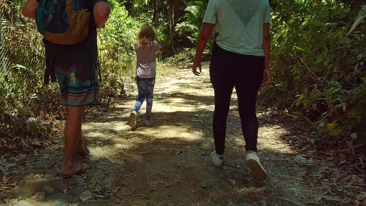 At 0:02, the child and two adults continue away along the leafy dirt trail in San Pancho, with bright sunlight on the path and thick vegetation at both sides.