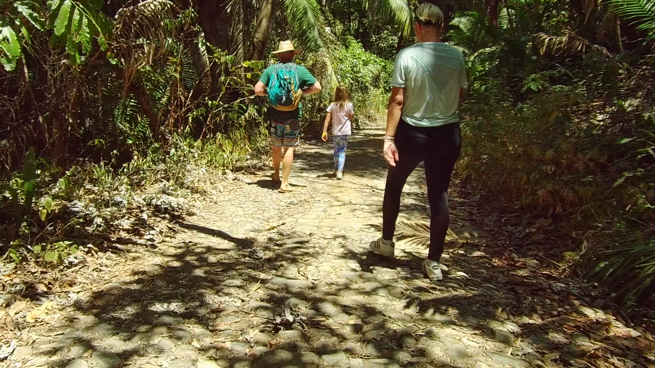 Three people walk away along a narrow shaded dirt path in San Pancho, Nayarit, Mexico, with dense green foliage around the trail.