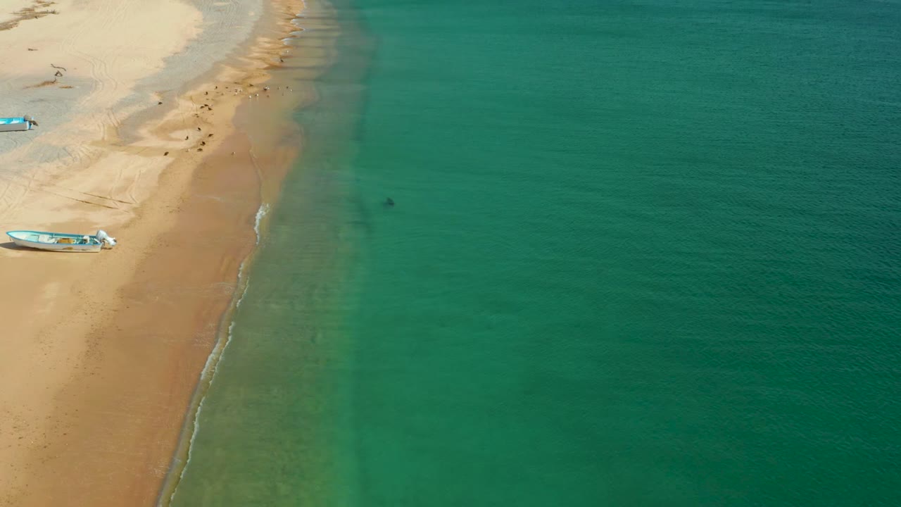 Aerial view over Los Frailes, Los Cabos, with tan sand on the left, turquoise ocean on the right, a small boat partly visible near the shore, and no visible people.