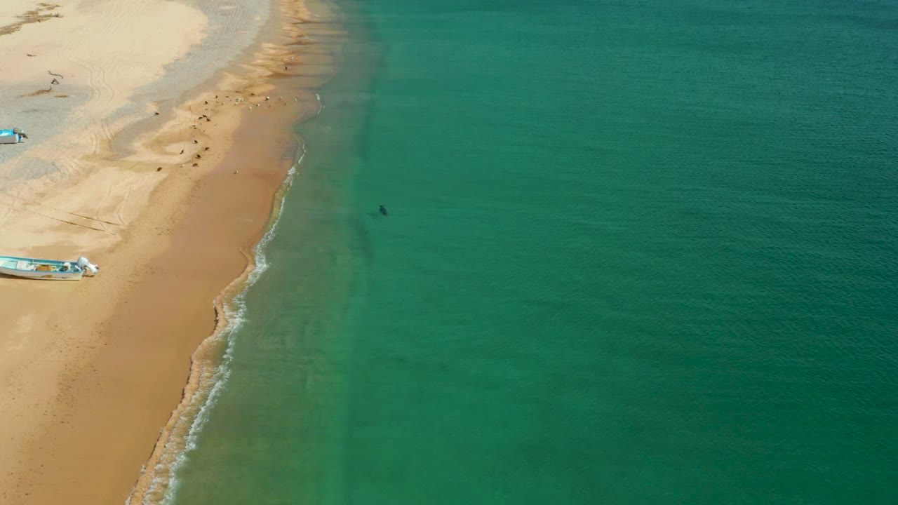 High view of the Los Frailes coast with open turquoise water dominating the right side, pale sand on the left, small boats partly visible, and no visible people.