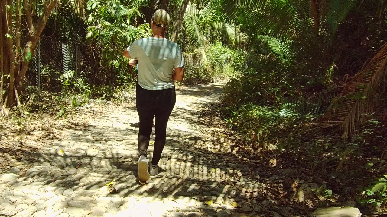 An adult in a pale shirt and dark leggings walks away on a dappled forest trail in San Pancho, Nayarit, Mexico, with dense greenery on both sides.