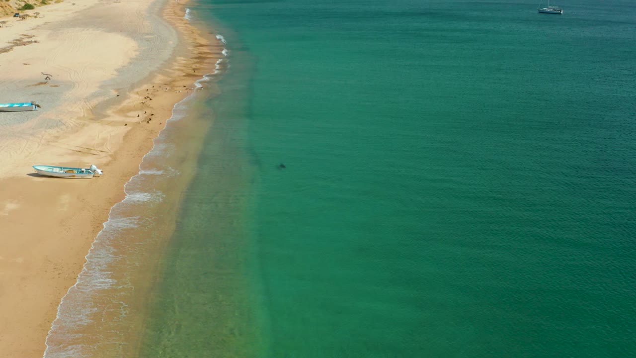 Aerial view in Los Frailes, Los Cabos, Baja California Sur showing the beach running diagonally along the left, a small boat on the sand, faint wave foam, and broad open turquoise water.
