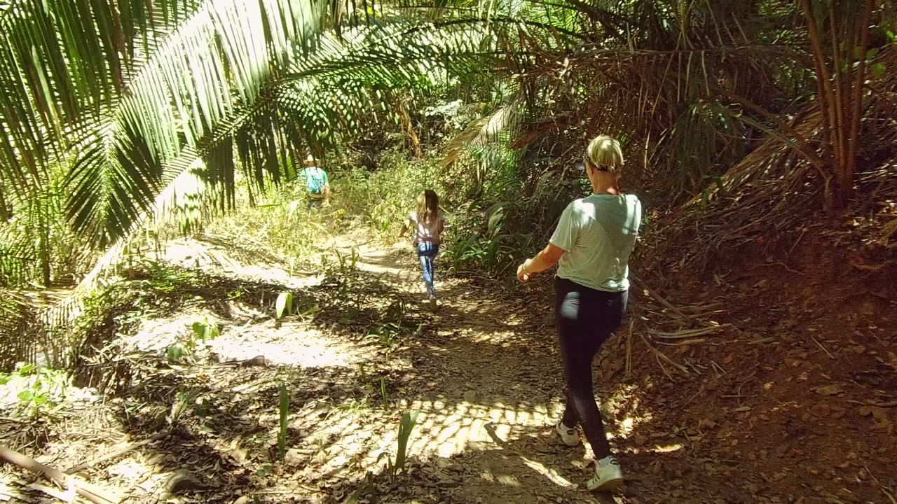 A woman in a light shirt walks behind another person on the San Pancho dirt path, with dense vegetation and palm fronds enclosing the trail.