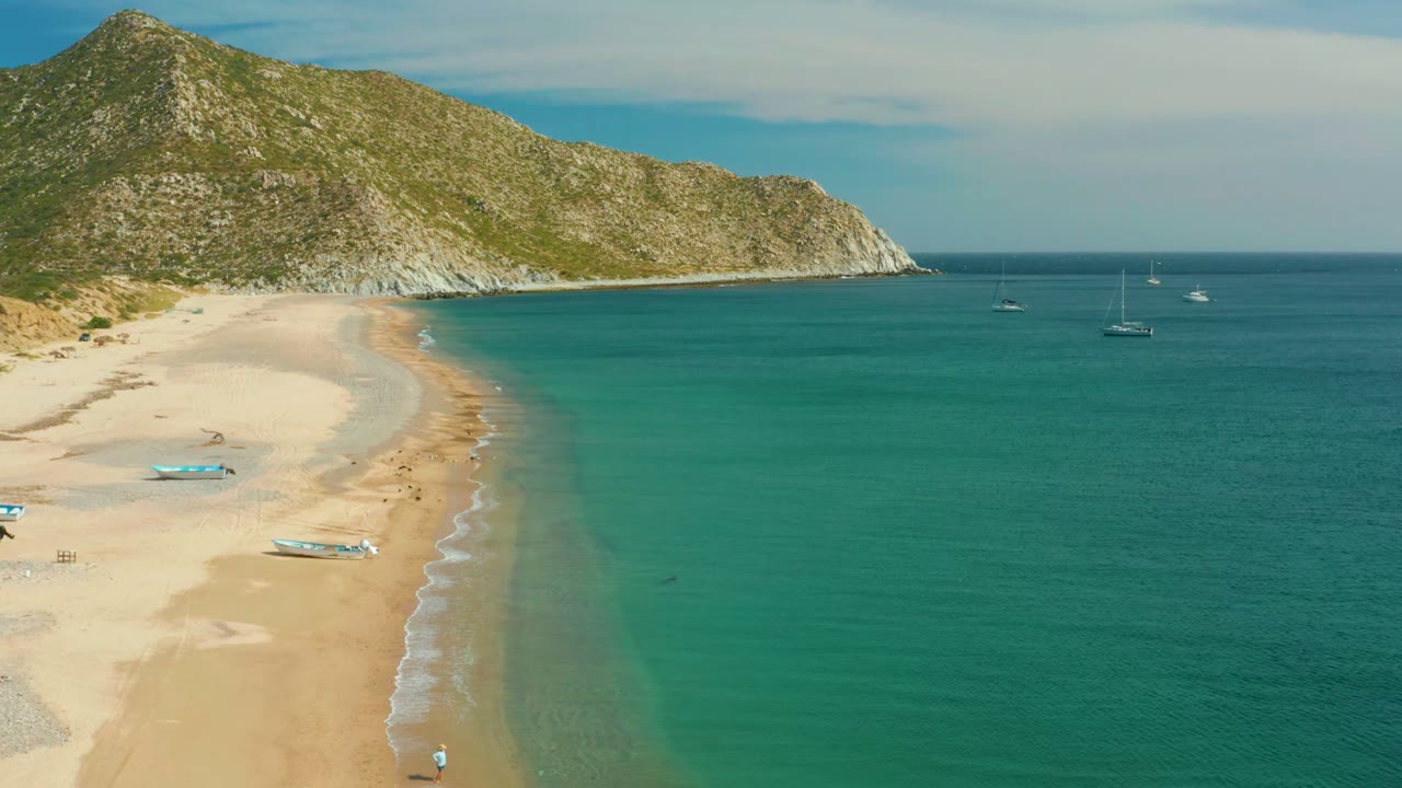 Aerial frame of Los Frailes beach showing the curved sandy cove, turquoise water, rocky green hillside, several anchored boats offshore, and tiny people visible near the shore.