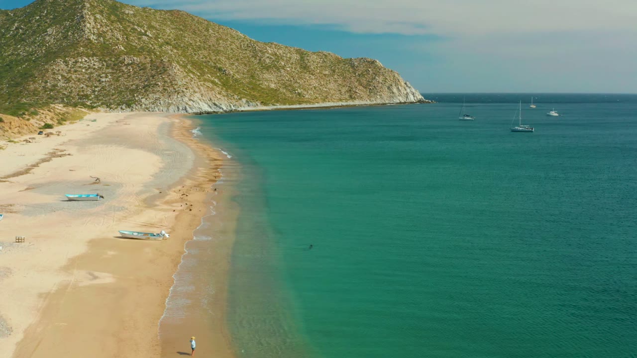 Aerial view over Los Frailes with the beach curving along the left side, clear green-blue shallows at center, anchored sailboats to the right, and small people visible on the sand.