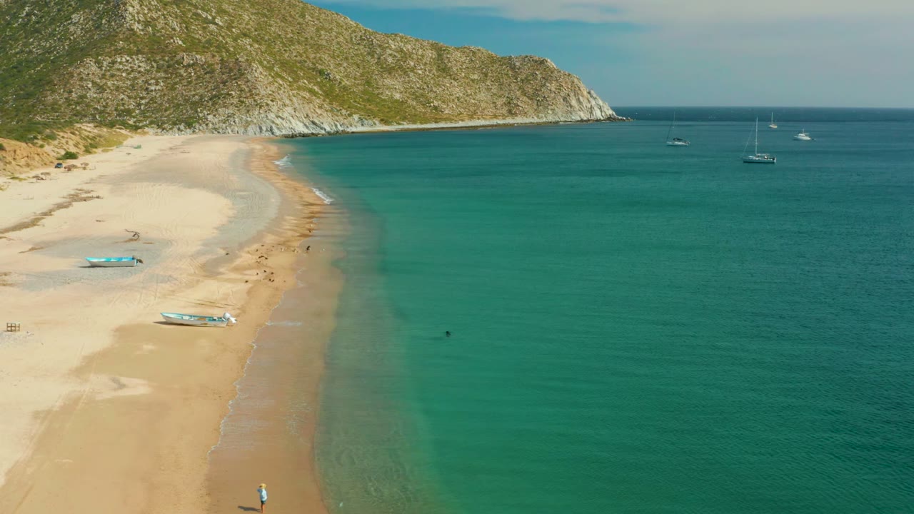 Wide Los Frailes aerial frame with more open turquoise water on the right, the sandy beach and small boats on the left, a hillside behind the cove, and tiny people visible near the shoreline.