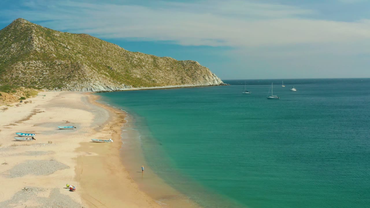 Aerial frame from Los Frailes Los Cabos showing a wide sandy curve, turquoise shallows, small boats on the beach, anchored sailboats offshore, a rocky green hillside, and no clearly visible people.