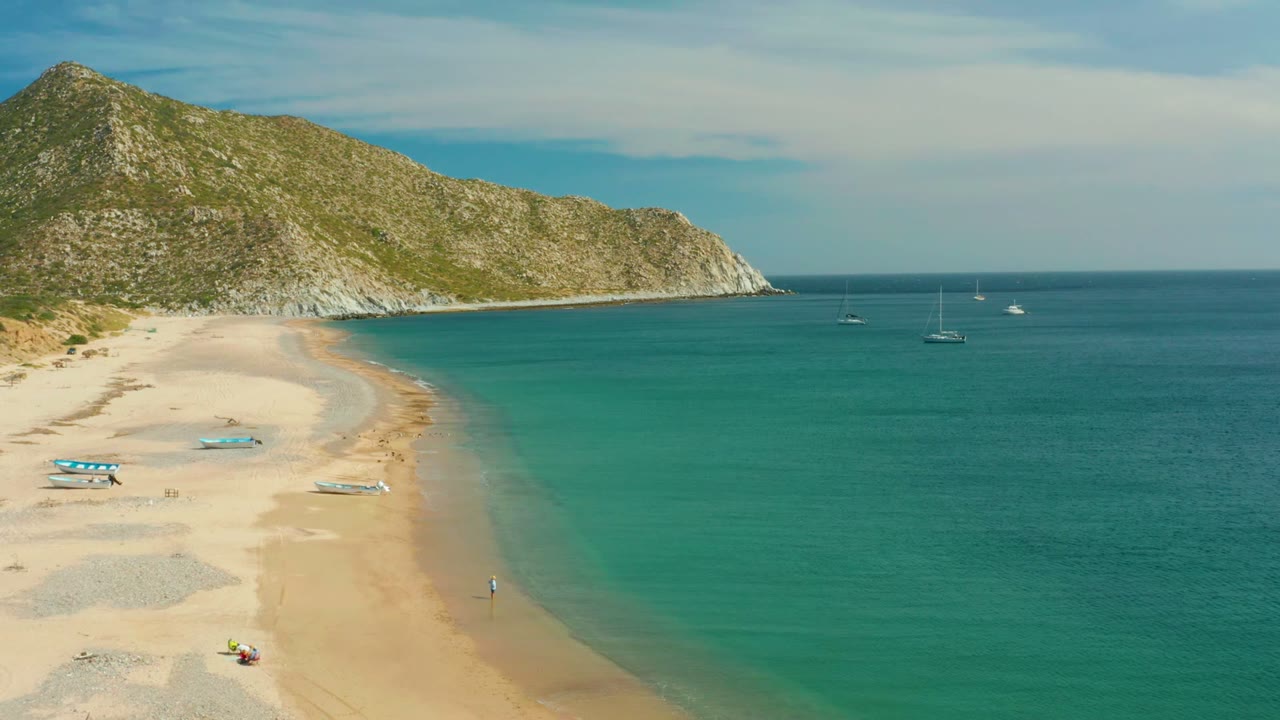Curving beach and shallow turquoise water with anchored boats offshore.