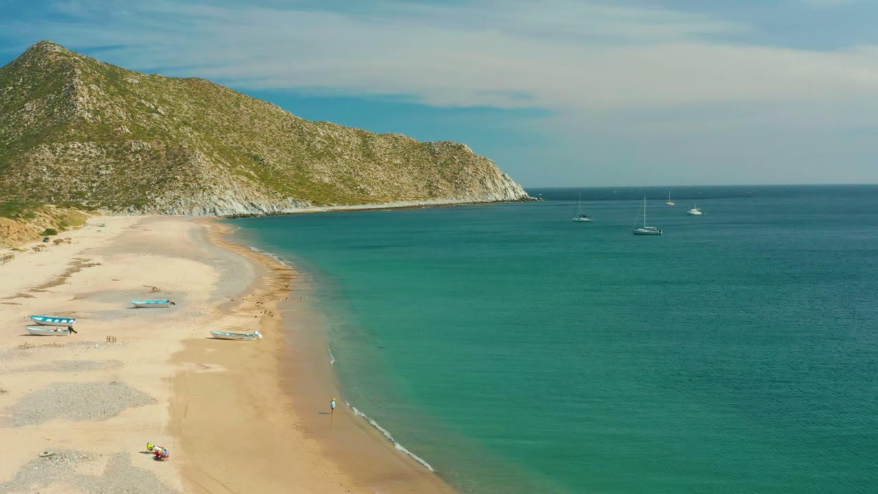 A wide Los Frailes Los Cabos beach frame shows the surf line beginning to brighten at the lower right, with turquoise water, pale sand, scattered small boats, offshore sailboats, hillside, and no clearly visible people.