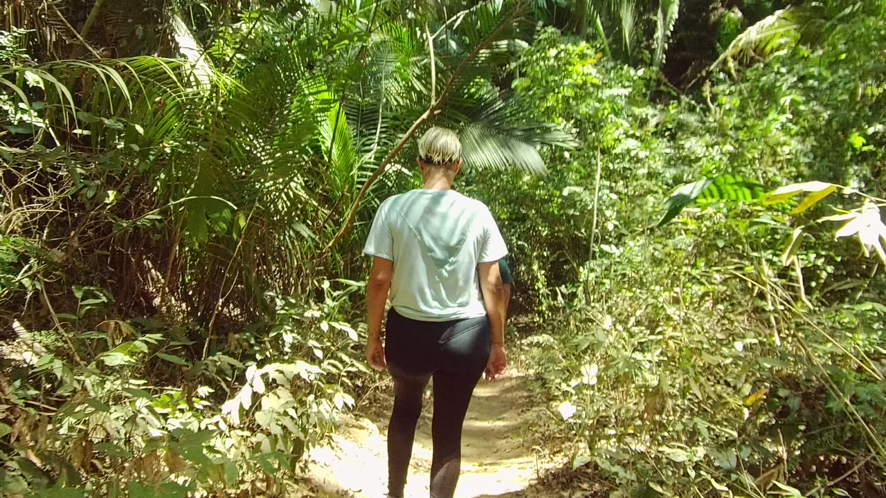 A person in a pale shirt and dark leggings walks away from camera on a narrow dirt trail in San Pancho, Nayarit, surrounded by dense green foliage.