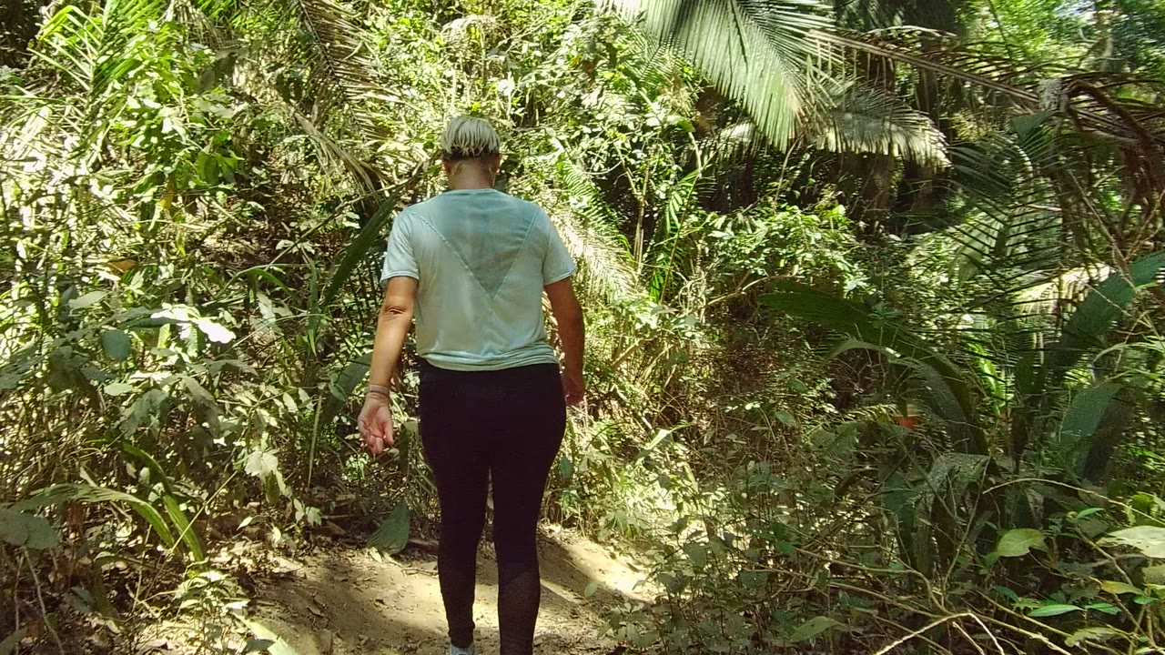A person in a pale blue shirt walks away from camera on a narrow dirt trail through dense green vegetation in San Pancho, Nayarit, Mexico.