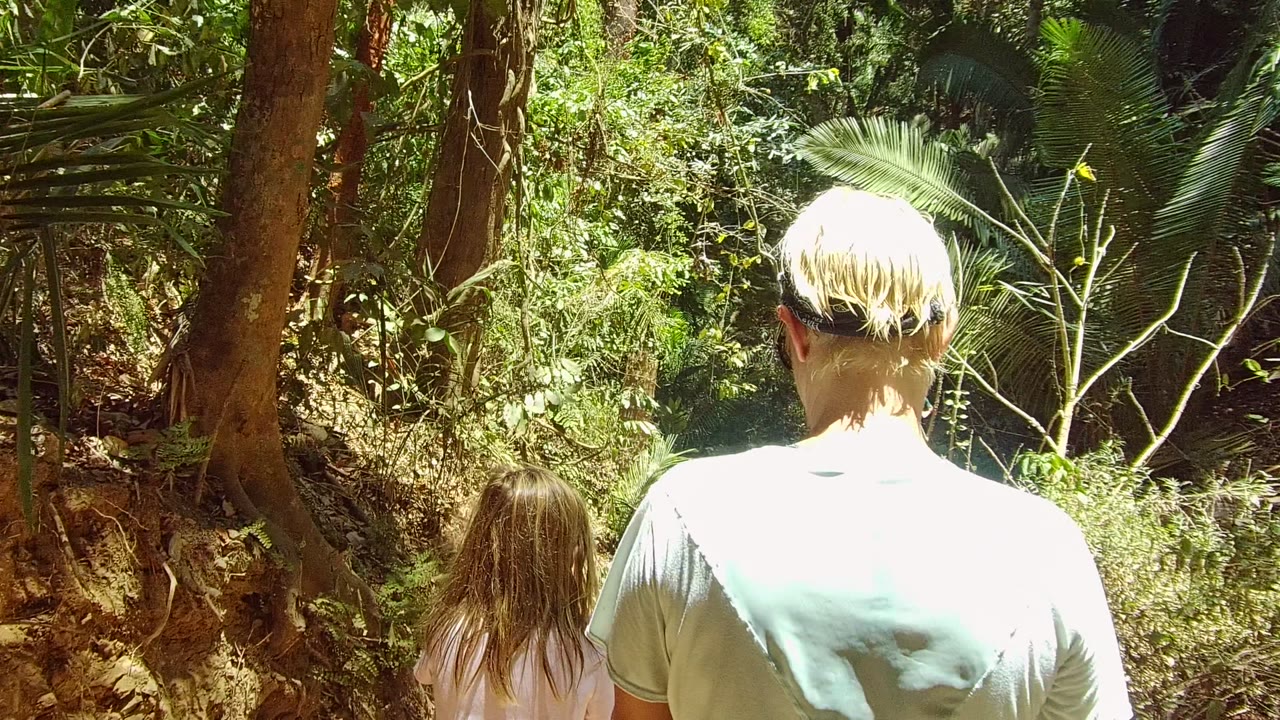 In San Pancho, Nayarit, the adult fills the right foreground while the child walks just ahead on the shaded dirt trail through thick vegetation.