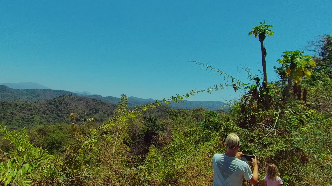 In San Pancho, Nayarit, a blond adult holding a phone and a child are partly visible from behind below dense green brush, with rolling forested hills and a clear blue sky beyond.
