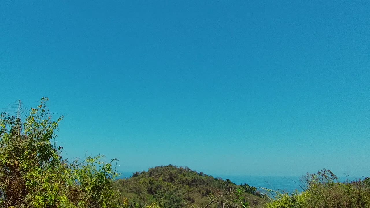 A forested hill rises behind foreground brush in San Pancho, Nayarit, with distant blue water on the horizon and no visible people.
